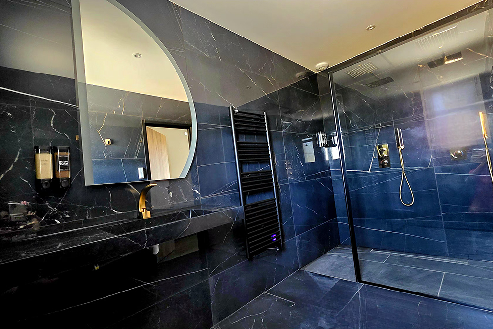 Luxurious black marble bathroom with a round mirror, gold faucet, black tiled walls, and a glass-enclosed shower.