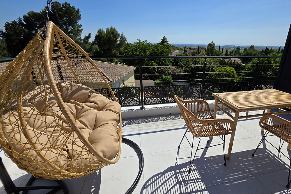 Outdoor balcony with a woven hanging chair, beige cushions, and a wooden table with chairs, overlooking trees and rooftops under a clear blue sky.