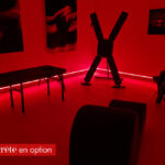 Red-lit private play room with BDSM equipment, including a large X-frame rack, and padded tables/benches in the foreground.