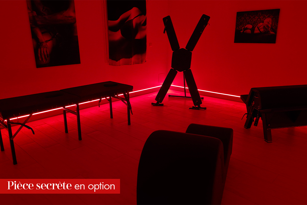 Red-lit private play room with BDSM equipment, including a large X-frame rack, and padded tables/benches in the foreground.