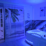 White jacuzzi tub with built-in seating under blue lighting in a spa room, beside a beach mural and open French doors to a tropical scene.
