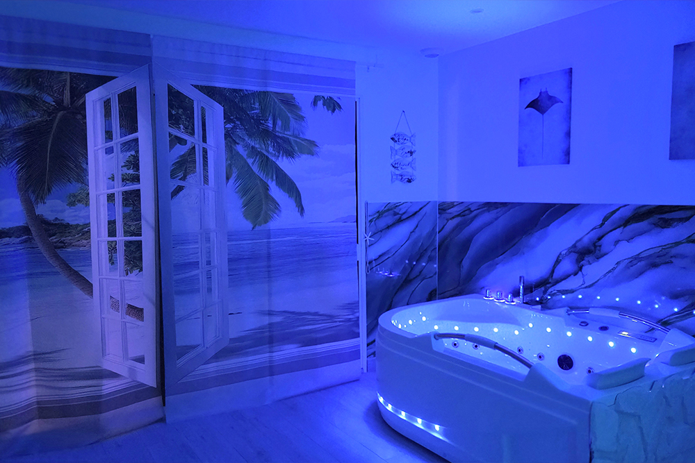White jacuzzi tub with built-in seating under blue lighting in a spa room, beside a beach mural and open French doors to a tropical scene.