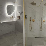 Modern marble bathroom with a glass shower enclosure, two gold rainfall showers, handhelds, and a backlit oval mirror above a curved sink for a cohesive, luxe look.