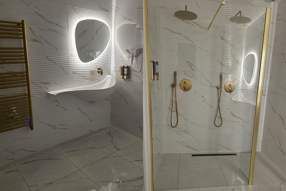 Modern marble bathroom with a glass shower enclosure, two gold rainfall showers, handhelds, and a backlit oval mirror above a curved sink for a cohesive, luxe look.