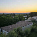 Sunset over a village with tiled-roof houses and dense green trees in the foreground.