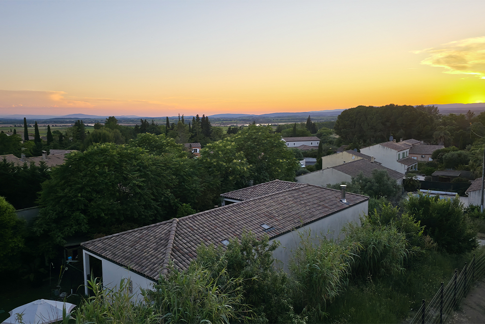 Sunset over a village with tiled-roof houses and dense green trees in the foreground.