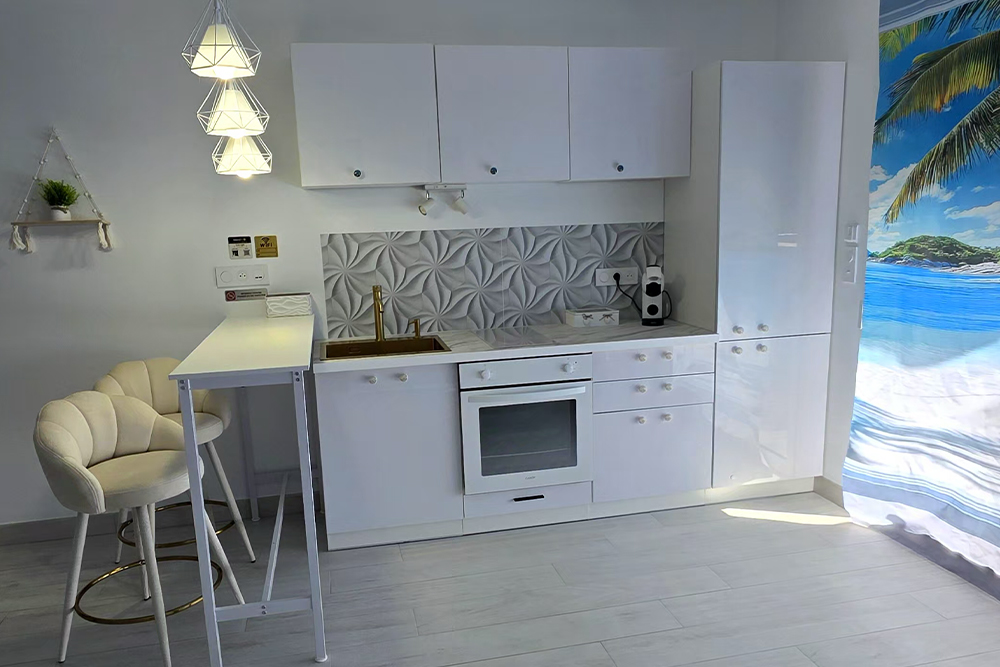 White modern kitchen with a bar-height table and two tufted stools, geometric tiled backsplash, and a tropical beach mural on the right.