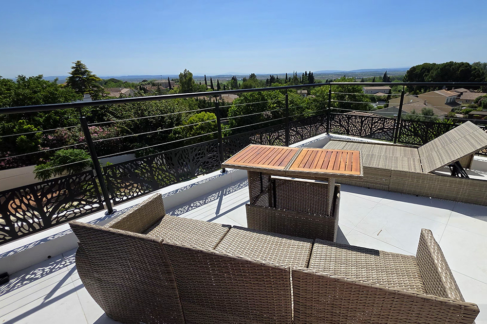 Rooftop terrace with wicker seating and a wooden table, overlooking a suburban neighborhood and distant hills on a sunny day.