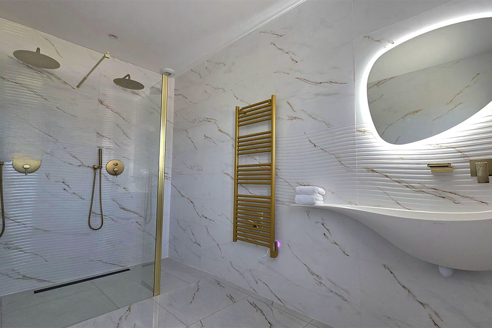 Modern marble bathroom with a glass shower enclosure and gold fixtures, a wall-mounted towel warmer, and a curved sink beneath an illuminated oval mirror.