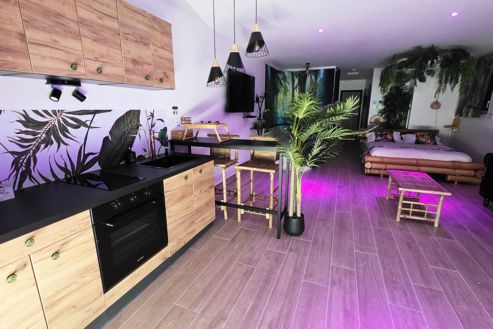 Open-plan tropical living space with a wooden kitchen, bar seating, and a bed area in the background under purple lighting, plus large potted plants and a leaf mural on the wall.