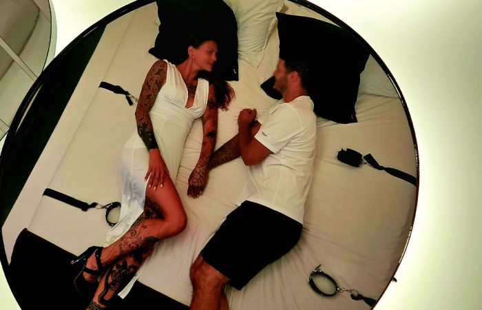 Tattooed couple lying on a bed inside a circular lighting setup, facing each other in white outfits with bondage straps nearby.